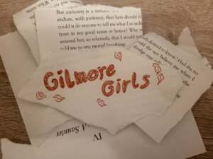 Viele Papierschnipsel auf einem Holztisch, die aus einem Buch rausgerissen wurden. Auf dem Schnipsel in der Mitte steht "Gilmore Girls" in roter Farbe. Daneben sind Zeichnungen von Blättern in der gleichen Farbe.
